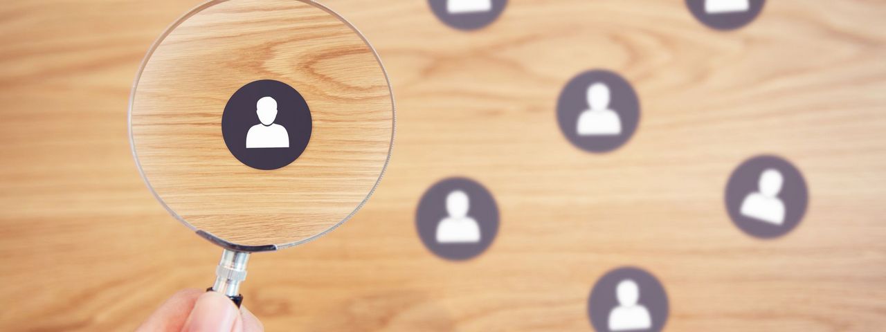 Aus eienr Gruppe symbolosiert dargestellter Menschen wird eine Person durch eine Lupe hervorgehoben